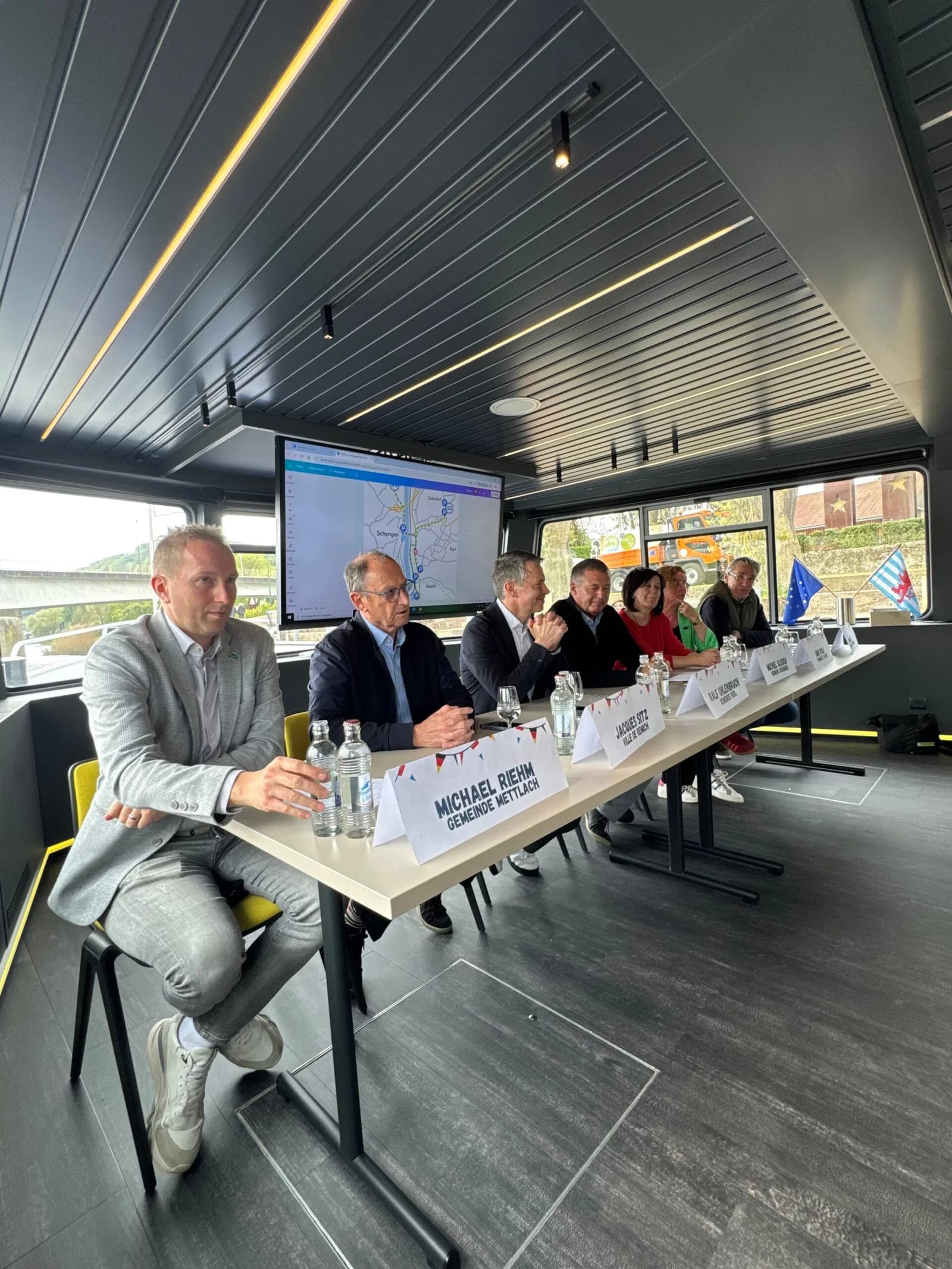 Des personnes assises à une table de conférence face à des micros et à un écran, dans une salle aux larges fenêtres.