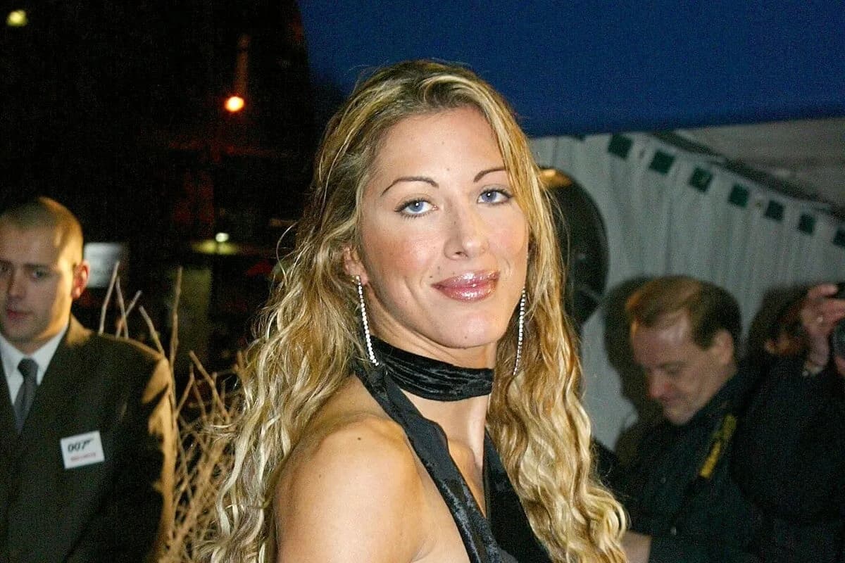 Une femme aux longs cheveux blonds bouclés sourit à l'objectif, de nuit, devant d'autres personnes.