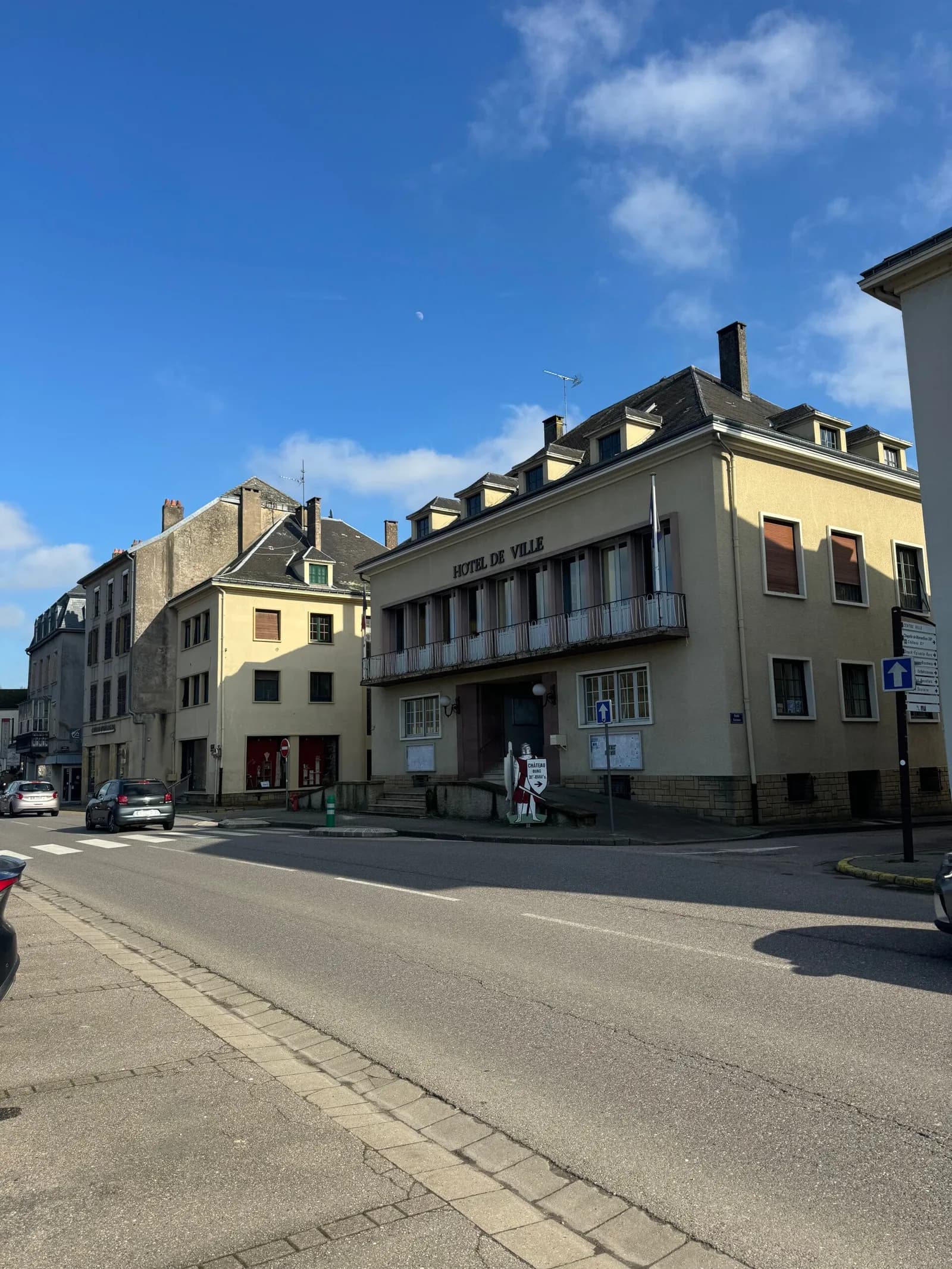 Bâtiment portant l'inscription « Hôtel de ville », avec un balcon et un drapeau, dans une rue ensoleillée.