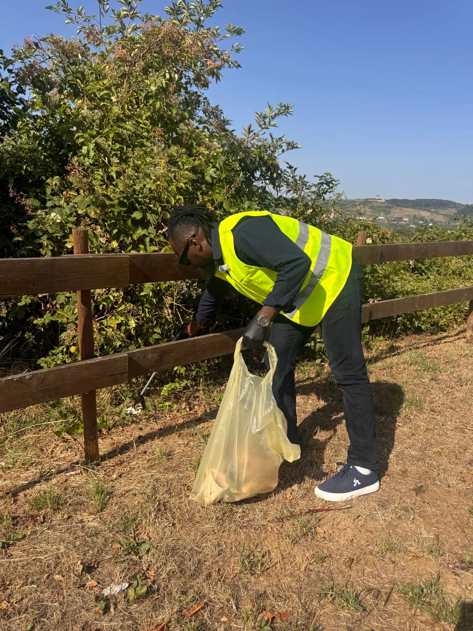 Une personne en gilet jaune remplit un sac de déchets près d'une barrière en bois, au milieu de buissons.