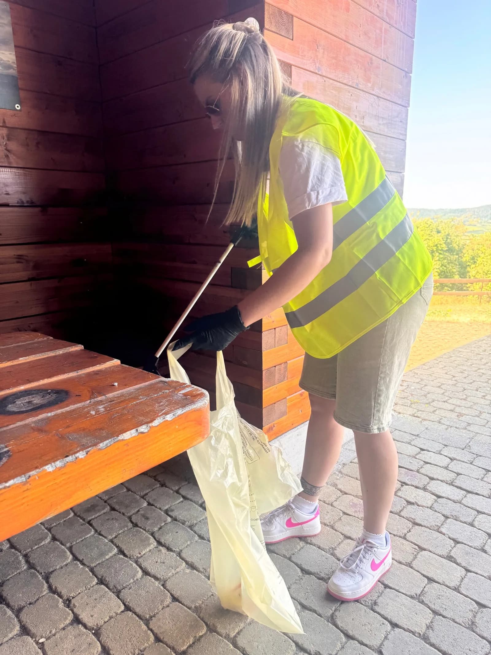 Une femme en gilet jaune tient un sac de déchets et une pince devant un abri en bois.