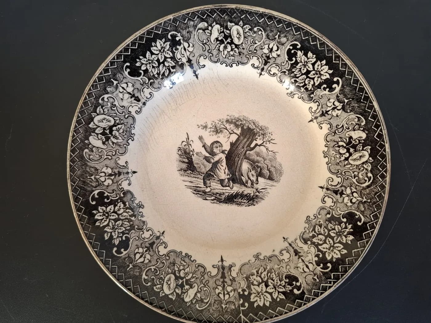 Assiette à décor imprimé noir : au centre, un enfant fuit un sanglier caché derrière un arbre ; large bordure de fleurs et d'arabesques.