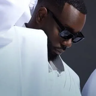 Un homme en tenue blanche et lunettes de soleil, tête baissée.