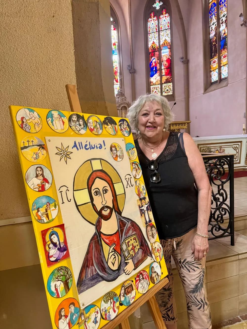 Anne-Marie Garandeau lächelt in einer Kirche mit bunten Fenstern neben einem großen, farbenfrohen Christusbild, umgeben von gemalten Medaillons.