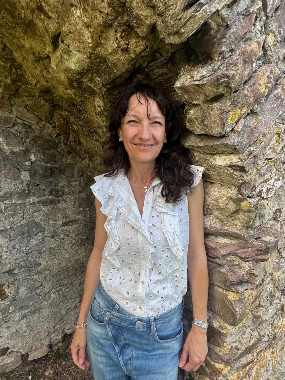 Frédérique Wehr lächelt in weißer Bluse, an eine alte Steinmauer gelehnt.