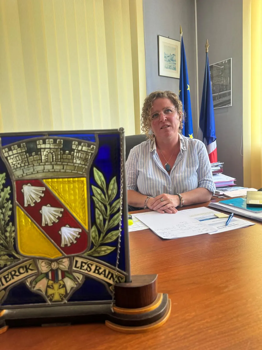 Helen Hammond an ihrem Schreibtisch im Rathaus vor der Europa- und der Frankreichflagge, im Vordergrund ein Glasbild mit dem Wappen von Sierck-les-Bains.